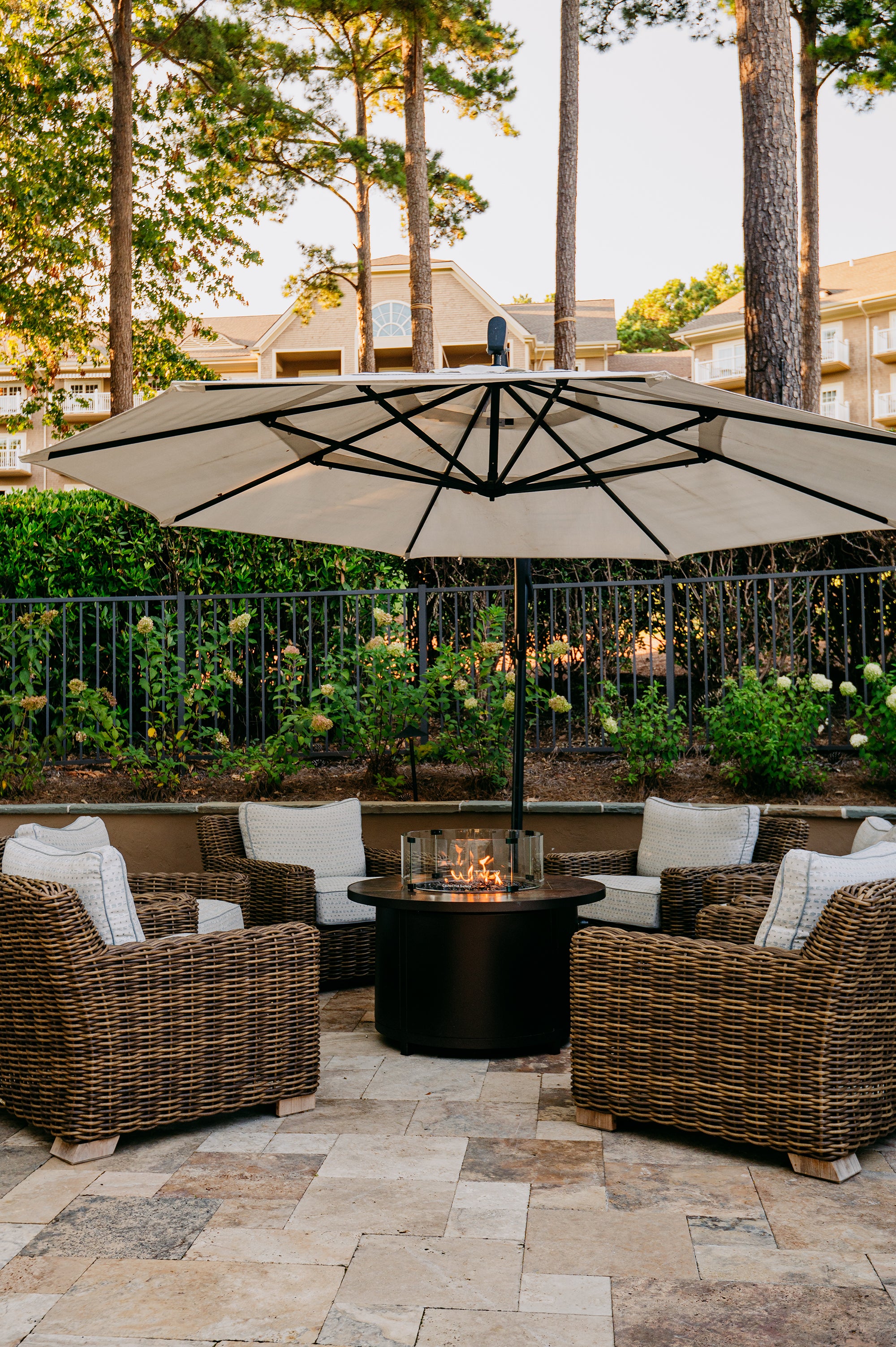 Outdoor patio with wicker chairs, fire pit, and large umbrella under trees.