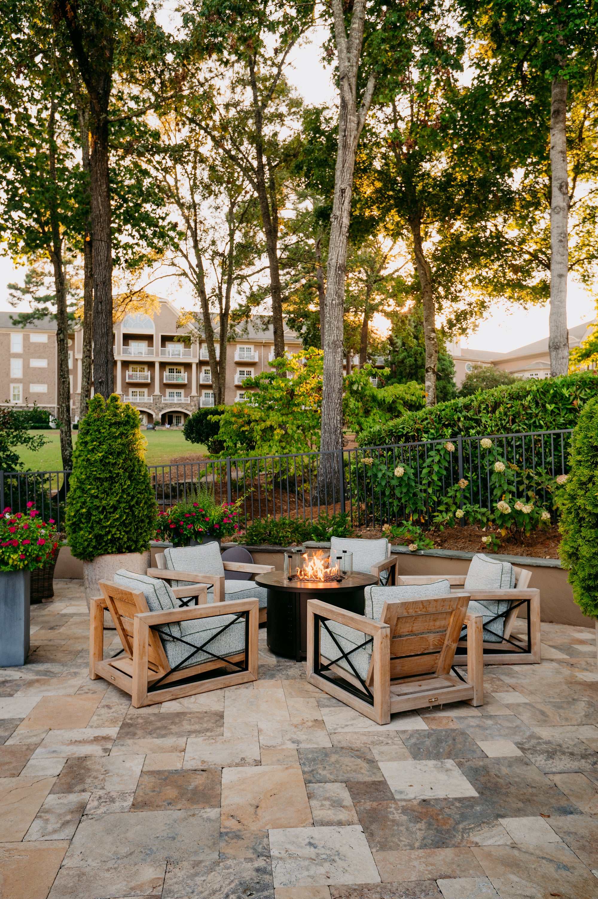 Outdoor patio setting with wooden chairs and a fire pit surrounded by trees and buildings.